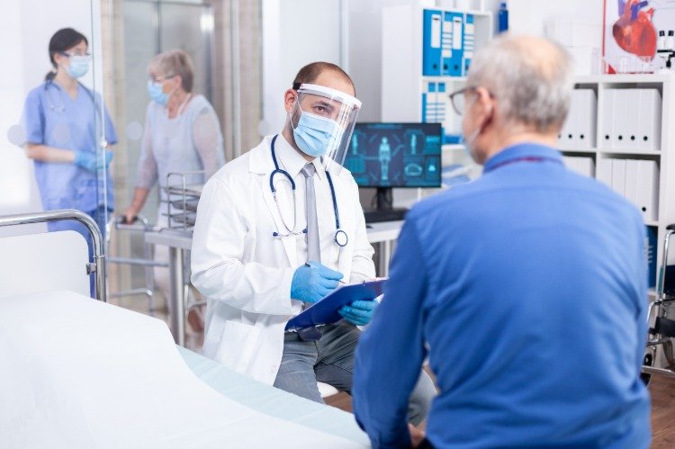 Lekár s ochranným štítom vyšetruje staršieho pacienta na príznaky covid 19