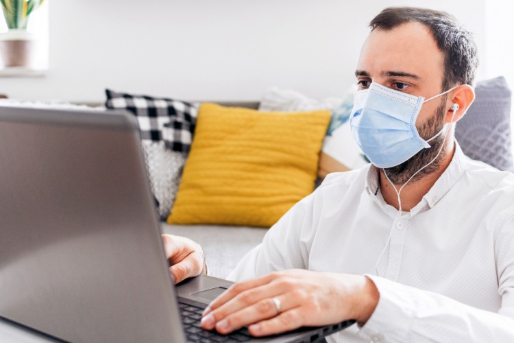mladý muž pracuje z domu na prenosnom počítači a doma nosí masku