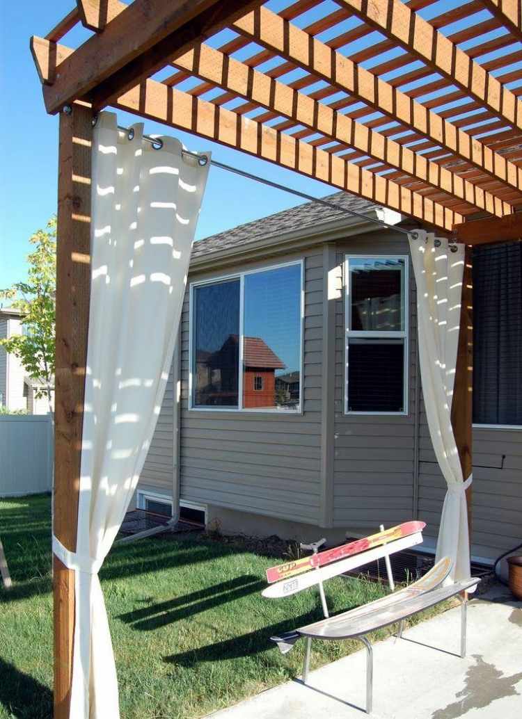 zaujímavá lavička zo starých lyží pod drevenou pergolou drevo-pergola-závesy-biela-slnečná clona-lavička-lyže-trávnik-dom-sivá