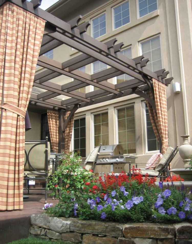 útulný vonkajší priestor s pergolou a pruhovanými závesmi drevo-pergola-závesy-záhradný gril-kvety-rastliny-tkanina-pruhované-okenné-záhradný nábytok