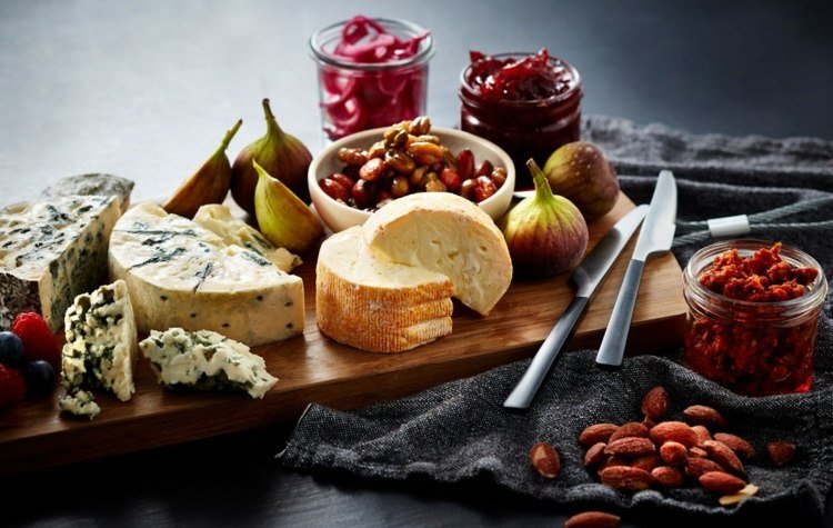 Rôzne druhy syrov s figami a orechmi na drevenej doske Podávanie syrového taniera nôž-kusy syra-obrus-fig-džem-murárske poháre-ovocno-paradajková pasta-dipová drevená doska