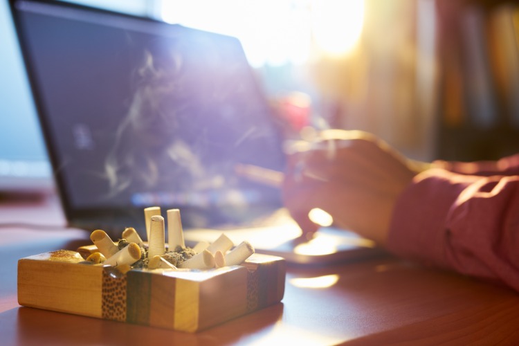 Popolník plný cigariet vedľa fajčiaceho muža pred prenosným počítačom