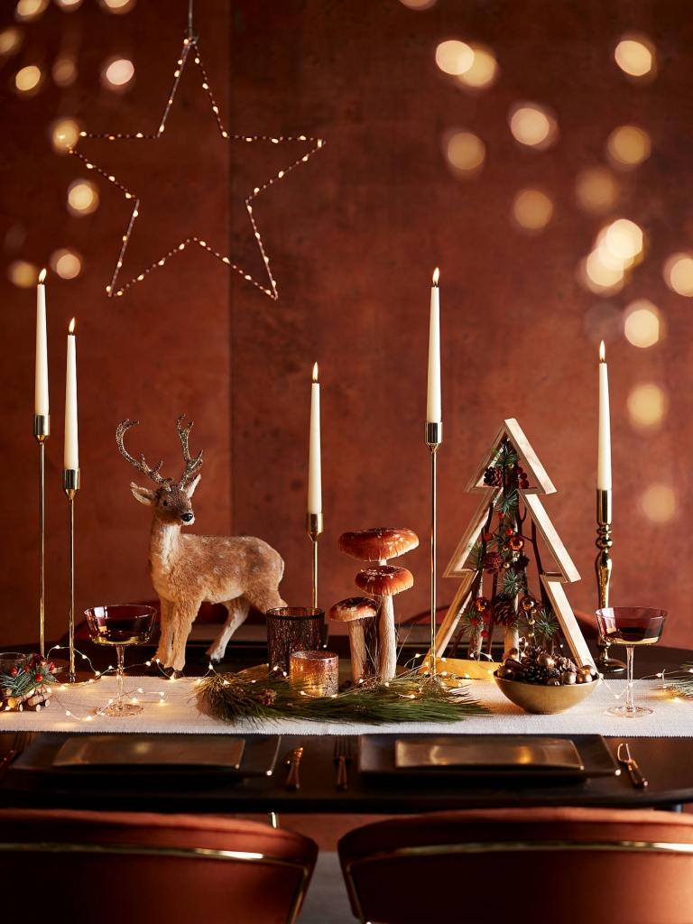 Nápady na slávnostné dekorácie stolu z medi a zlata s rozprávkovými svetlami ako akcentmi a ozdobnými figúrkami jeleňov a húb a vianočného stromčeka z dreva