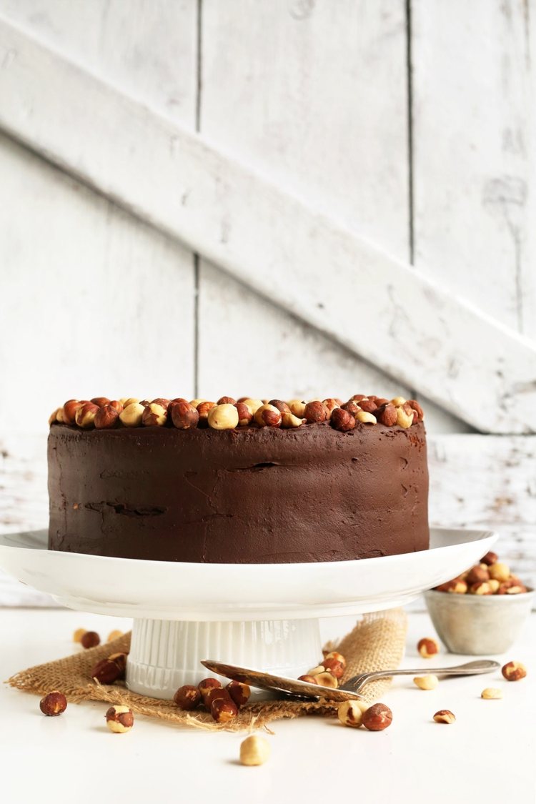 Vegánsky čokoládový koláč ozdobený lieskovými orieškami vegánska čokoládová torta lieskové oriešky-torta-čokoládová-poleva-torta-tanier-vrstvené-misa-torta server