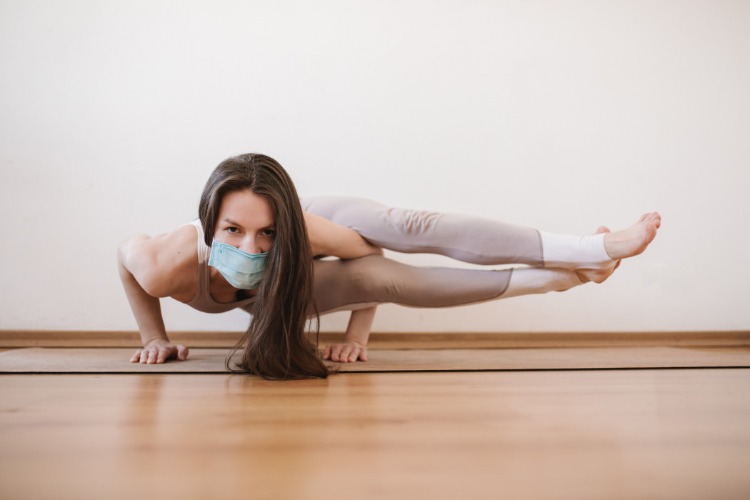 Mladá žena s tvárovou maskou cvičí doma dištančnú jogu a meditáciu